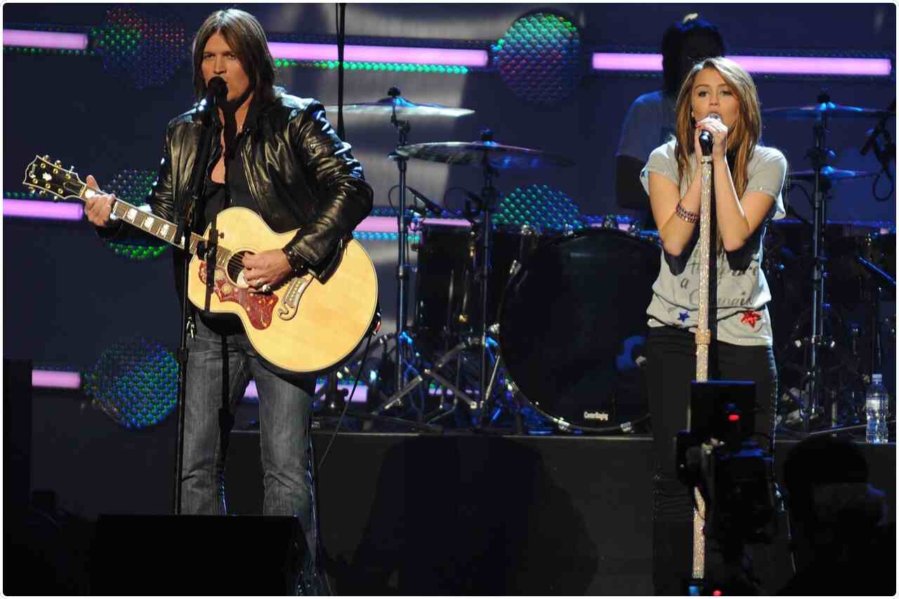 Billy Ray Cyrus and Miley Cyrus posing together with guiter in hand and singing in stage.